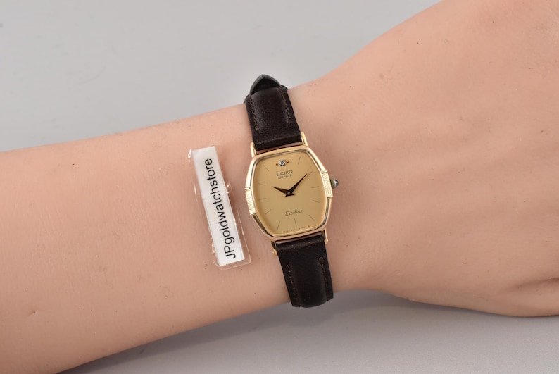 Puede incluir: Un reloj de pulsera Seiko Quartz con caja dorada en forma de tonel, esfera color champ&aacute;n y correa de cuero marr&oacute;n oscuro. La esfera presenta manecillas negras, un peque&ntilde;o diamante en la posici&oacute;n de las doce y el texto JPgoldwatchstore.
