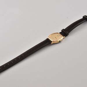 Puede incluir: Un reloj de pulsera con caja rectangular redondeada en tono dorado, esfera dorada a juego y una correa fina de cuero negro texturizado. El reloj incluye un cierre de hebilla en tono dorado y una peque&ntilde;a corona lateral para el ajuste.