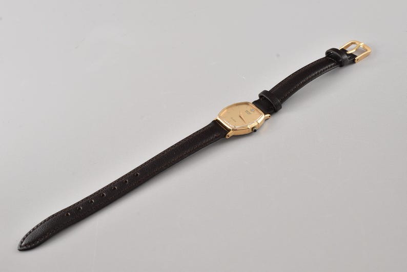 Puede incluir: Un reloj de pulsera con caja rectangular redondeada en tono dorado, esfera dorada a juego y una correa fina de cuero negro texturizado. El reloj incluye un cierre de hebilla en tono dorado y una peque&ntilde;a corona lateral para el ajuste.