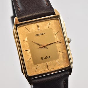 Puede incluir: Un reloj Seiko Dolce dorado con esfera rectangular y correa de cuero marrón. La esfera del reloj presenta una superficie dorada texturizada con marcadores y agujas negras. La palabra "Dolce" está impresa en la esfera.
