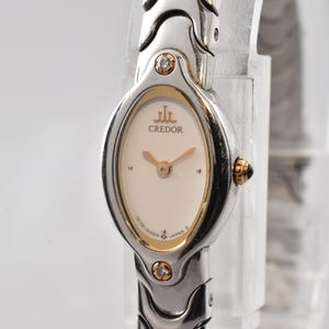 May include: A silver-tone wristwatch with an oval-shaped case and a cream-colored dial. The watch features gold-tone hands, a gold-tone bezel, and two small clear stones set at the top and bottom of the case. The word CREDOR is printed on the dial.
