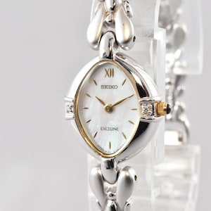 Könnte beinhalten: Eine Seiko Exceline Armbanduhr mit einem silberfarbenen Metallgliederarmband und einem perlmuttfarbenen Zifferblatt. Das ovale Gehäuse hat goldfarbene Akzente, eine römische Ziffer auf der Zwölf-Uhr-Position und kleine klare Steine.