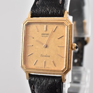 Op de afbeelding: Een Seiko Quartz Exceline polshorloge met een rechthoekige goudkleurige kast en een getextureerde lichtbruine wijzerplaat. Het horloge heeft dunne goudkleurige uurmarkeringen, bijpassende wijzers en een zwarte leren band met zichtbare stiksels langs de randen.
