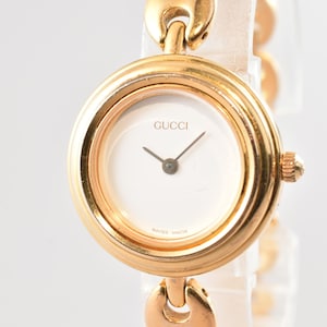 May include: A gold-tone wristwatch with a round case and a white dial. The dial features the Gucci logo at the top and the text Swiss Made at the bottom. It has two thin hands and a textured crown on the side. The band consists of gold-tone links.