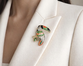 May include: A toucan-shaped brooch featuring gold-tone metal, vibrant enamel in shades of green, orange, and black, and clear crystal accents. The bird is depicted perched on a branch, showcasing detailed plumage patterns and a curved beak.