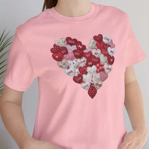 Peut inclure: Un t-shirt rose clair orné d'un motif en forme de cœur composé de cœurs en tissu rouges, blancs et roses. Certains cœurs affichent des mots comme "LOVE" et "XOXO". Le t-shirt est à col rond et à manches courtes.