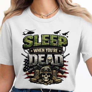 May include: White t-shirt with a military-themed graphic. The design features the text "SLEEP WHEN YOU'RE DEAD" in camouflage lettering, along with a skull, guns, boots, and the American flag. Helicopters and fighter jets are also depicted.