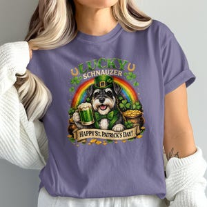 May include: A purple t-shirt featuring a cartoon Schnauzer wearing a green hat and bow tie, holding a beer mug. The design includes a rainbow, shamrocks, and the text "Lucky Schnauzer" and "Happy St. Patrick's Day!"
