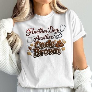 Puede incluir: Camiseta blanca con la frase "Another Day, Another Code Brown". El diseño incluye un gorro de enfermera, un estetoscopio, papel higiénico, una jeringa y un emoji de caca sonriente. El texto y los gráficos son marrones y rojos.