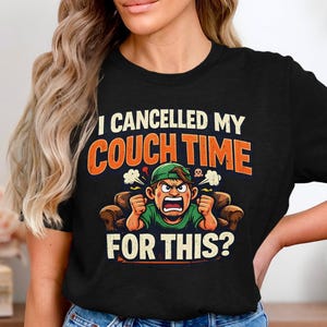 May include: Black t-shirt with the text "I Cancelled My Couch Time For This?" in white and orange. The graphic features an angry cartoon character in a green shirt and hat, sitting on a brown couch.