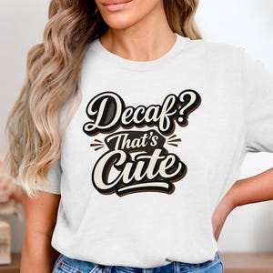 May include: White t-shirt featuring the phrase "Decaf? That's Cute" in a black and brown retro font. A casual style t-shirt suitable for everyday wear.