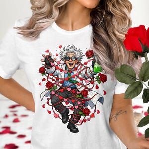 May include: White t-shirt featuring a cartoon scientist entangled in heart-shaped garland and roses. The scientist holds a green liquid in a flask. A large red rose is held in the hand.