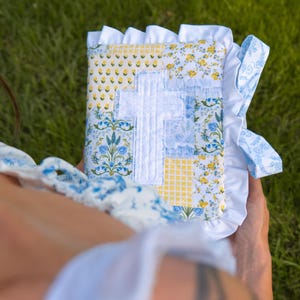 May include: A square fabric book cover featuring a patchwork design of yellow and blue floral and gingham patterns. A white fabric cross is appliqued in the center. The edges are finished with a white ruffled trim and a blue floral ribbon tie.