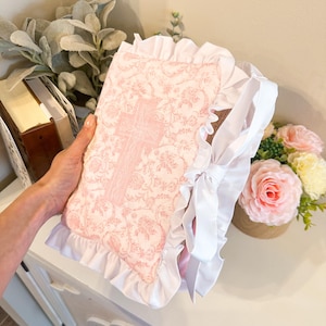 以下が含まれることがあります： ピンクと白の布で覆われた本で、十字のデザイン。本は花柄で、白いフリルと白いリボンが付いています。表紙は薄いピンク色です。