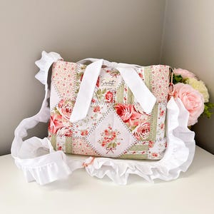 May include: A patchwork crossbody bag with a floral pattern in pink, red, and green. The bag has a white ruffled trim and a white strap. The text "Sewtist" is visible on the front of the bag.