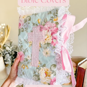 May include: A quilted fabric Bible cover featuring a light blue background with a floral pattern of pink and yellow roses. The cover is adorned with a central pink fabric cross, white lace trim along the edges, and a pink ribbon tie. Text reads Bible Covers.