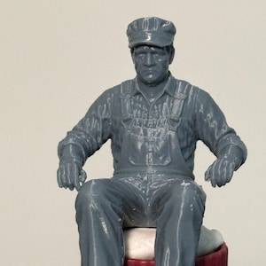 May include: A gray figurine of a seated man wearing a cap, overalls, and a long-sleeved shirt. The figure is seated on a red and black cylindrical base. The man's arms are resting on his legs.
