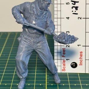 May include: A gray scale model of a worker in overalls and a cap, holding a shovel filled with coal. The model is standing next to a ruler with the text "Scale Feet | Inches" and the number "1:24".