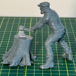 May include: A gray figurine of a lumberjack swinging an axe at a tree stump. The lumberjack wears a cap, work clothes, and boots. The axe is raised above the stump, ready to strike. The scene is set on a green grid.