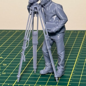 May include: A gray miniature figurine of a man using a surveying instrument on a tripod. The figure is wearing a jacket and pants, holding a notepad. The tripod has adjustable legs. The background is a green cutting mat.