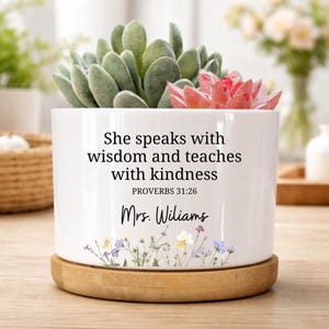 May include: White ceramic planter with a succulent arrangement. The planter has the text "She speaks with wisdom and teaches with kindness" and "Mrs. Wiliams". The planter sits on a round wooden base.
