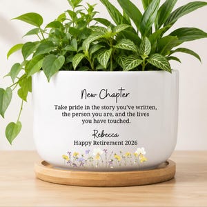 May include: White ceramic planter with the text "New Chapter" and a personalized message. The planter is filled with green plants and sits on a wooden saucer. The bottom of the planter has a floral design.