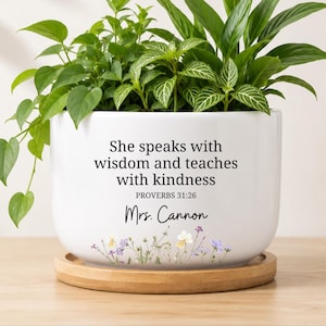 May include: White ceramic planter with the text "She speaks with wisdom and teaches with kindness" and "PROVERBS 31:26". The planter has a floral design and is filled with green plants. It sits on a wooden saucer.