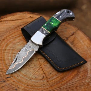 Peut inclure: Un couteau de poche pliant avec une lame à motifs et un manche vert et noir. Le couteau est ouvert, posé sur une surface en bois à côté d'un étui en cuir noir. La lame a un accent de couleur cuivre.