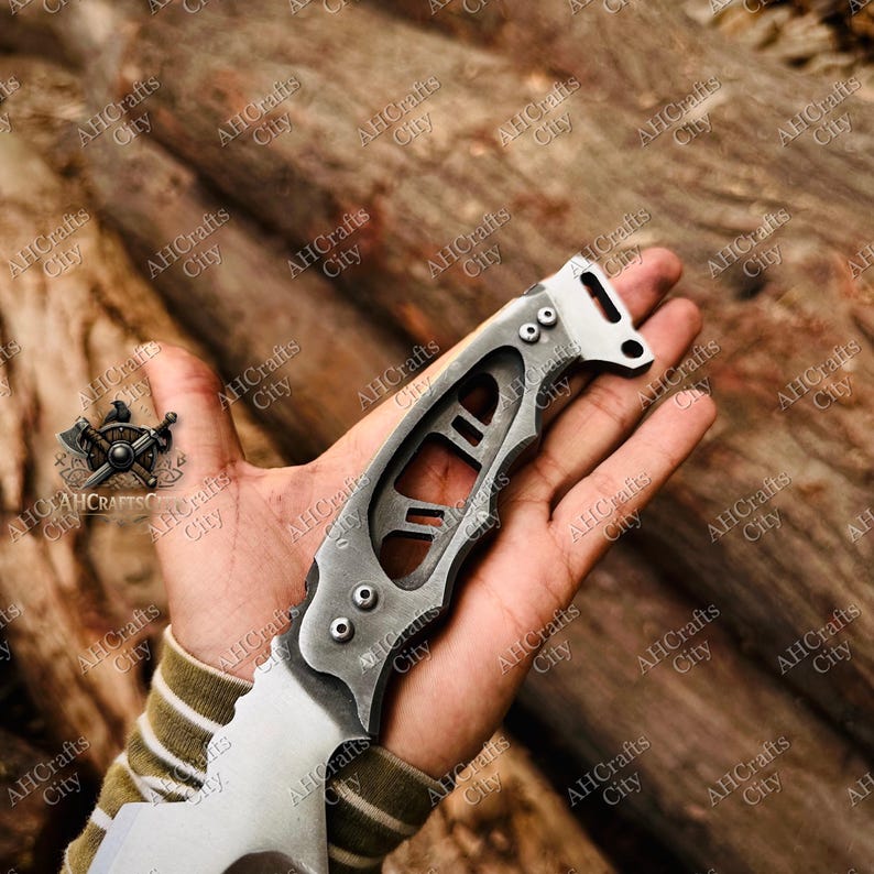 Peut inclure: Un couteau pliant argent&eacute; et gris avec une lame partiellement dentel&eacute;e. Le manche a un design ouvert unique avec trois rivets et un trou &agrave; l'extr&eacute;mit&eacute;. Le couteau est tenu dans une main sur un fond en bois.