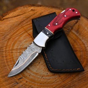Peut inclure: Couteau pliant en acier de Damas avec un manche en bois rouge et un étui en cuir noir. La lame présente un motif décoratif et un accent cuivré. Le manche est orné d'incrustations de points blancs.