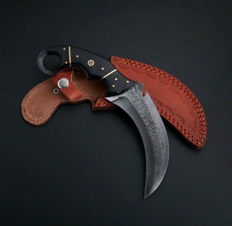 Peut inclure: Un couteau karambit incurv&eacute; avec un manche noir et une lame &agrave; motifs, reposant dans un &eacute;tui en cuir marron. La poign&eacute;e est orn&eacute;e d'accents dor&eacute;s. Le couteau est con&ccedil;u pour l'utilit&eacute; et l'autod&eacute;fense.