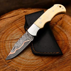 Peut inclure: Un couteau pliant avec un manche crème et une lame à motifs avec des accents cuivrés. Le couteau est posé sur une surface en bois à côté d'un étui en cuir noir. La lame présente un motif texturé unique.