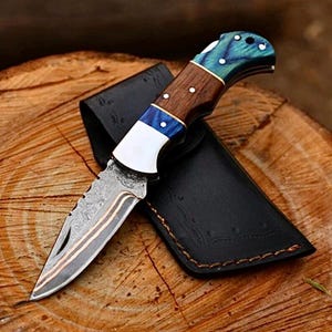 Peut inclure: Un couteau pliant avec une lame à motifs et un manche en bois avec des accents bleus et blancs. Le couteau est dans un étui en cuir noir, posé sur une surface en bois. La lame a un motif décoratif.