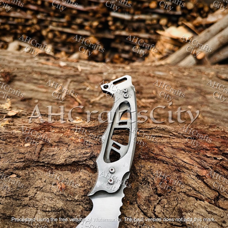 Peut inclure: Un couteau argent&eacute; avec une lame dentel&eacute;e et une poign&eacute;e au design unique, avec des d&eacute;coupes et des rivets. Le couteau est pr&eacute;sent&eacute; sur une surface textur&eacute;e et brune, probablement en bois. Le texte "AHCrafts City" est en filigrane.