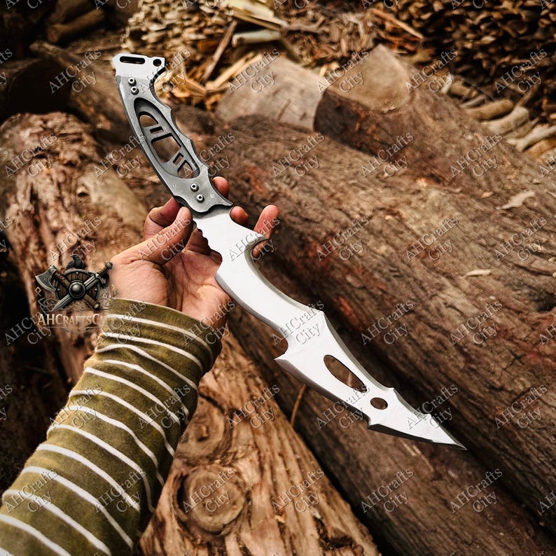 Peut inclure: Un couteau argent&eacute; et noir avec une lame incurv&eacute;e et un manche avec des motifs d&eacute;coup&eacute;s. Le couteau est tenu dans une main sur un fond de b&ucirc;ches de bois. La lame a une pointe ac&eacute;r&eacute;e.