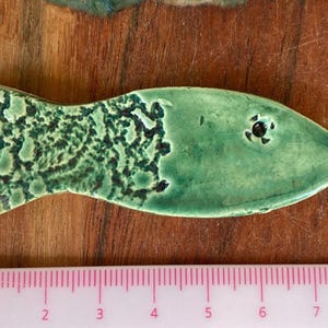 Könnte beinhalten: Handgefertigte Keramik-Fischfigur in Grüntönen. Der Fisch hat ein strukturiertes Muster auf seinem Körper und ein dunkles Auge. Ein rosa Lineal mit Zentimetermarkierungen befindet sich im Vordergrund.