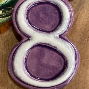 Pode incluir: Um número oito de cerâmica em tom roxo com contorno branco. O número é brilhante e parece ser feito à mão. O fundo é uma superfície de madeira com uma régua na parte inferior.