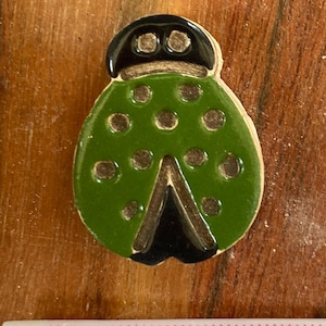 May include: A green ladybug-shaped decorative item with black accents. The ladybug has a glossy finish and features dark spots on its back. The item is approximately 1 inch long and is set against a wooden background.
