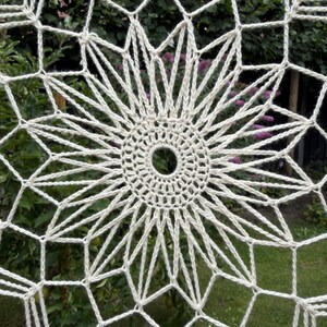 Op de afbeelding: Een gebroken wit gehaakt kleedje met een cirkelvormig midden met een klein gat, omringd door stralende stervormige punten en een geometrisch ruitpatroon dat zich uitstrekt tot aan de randen van het ronde textielstuk.