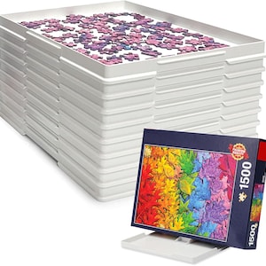 May include: A stack of white plastic sorting trays for jigsaw puzzles, with the top tray containing scattered purple and blue puzzle pieces. A vertical puzzle box featuring a colorful rainbow leaf design and the text 1500 is displayed in front.