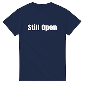 May include: A navy blue t-shirt with the words "Still Open" printed in bold white letters across the chest. The shirt is a classic crew neck style, suitable for casual wear.