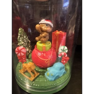 Op de afbeelding: Een kerst-sneeuwbol met een bruine beer met een kerstmuts op een rode cadeautas. Bevat een kerstboom, cadeaus, een schommelpaard, een blauwe auto en zuurstokken.