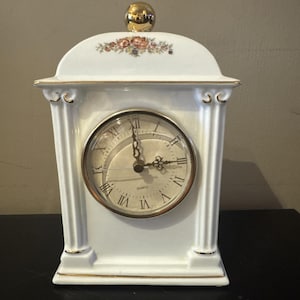 May include: White porcelain mantel clock with a gold-colored top and trim. The clock face has Roman numerals and a gold-colored bezel. Floral accents are on the top of the clock.