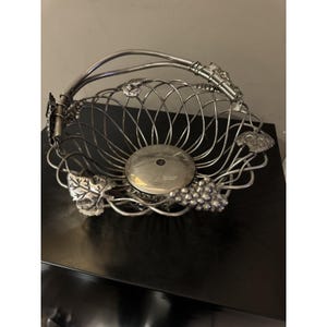 May include: Silver-toned wire fruit basket with a handle. The basket features a round base and is decorated with grape and leaf accents. The handle is arched and attached to the basket with decorative elements. The basket is designed to hold fruit.