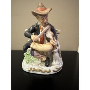 Peut inclure: Figurine en céramique d'un homme assis jouant du violon. L'homme porte un chapeau marron, une veste noire et une cravate rouge. La figurine est peinte avec des détails et repose sur une base avec un petit chien et du feuillage.