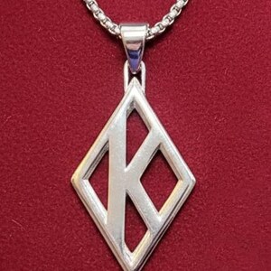 May include: A silver pendant necklace featuring a diamond-shaped pendant with a stylized letter "K" cut out. The pendant hangs from a silver chain against a red background. The necklace is a piece of jewelry.