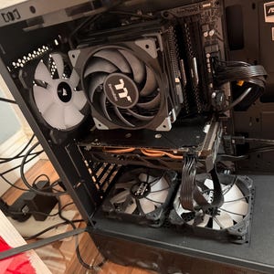 May include: Inside view of a computer case with various components. A large black and gray CPU cooler is centered, with a graphics card below. Several fans are visible, including a white Corsair fan and two others.