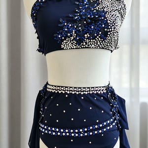 May include: A navy blue two-piece dance costume featuring a crop top and high-waisted bottoms. The top is adorned with blue and clear crystals and floral lace appliqués. The bottoms feature crystal-embellished waistbands and side drapes.