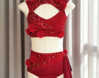 May include: A two-piece red dance costume featuring a high-neck crop top with a cutout, adorned with sequins and fabric flowers. The matching high-waisted briefs include a side-draped chiffon panel, also embellished with sequins and flowers.