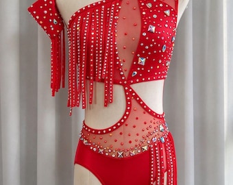 May include: A red one-shoulder leotard featuring sheer mesh cutouts, long red fringe accents, and scattered iridescent rhinestones and square crystals. The garment includes a matching detachable sleeve piece and is displayed on a mannequin.